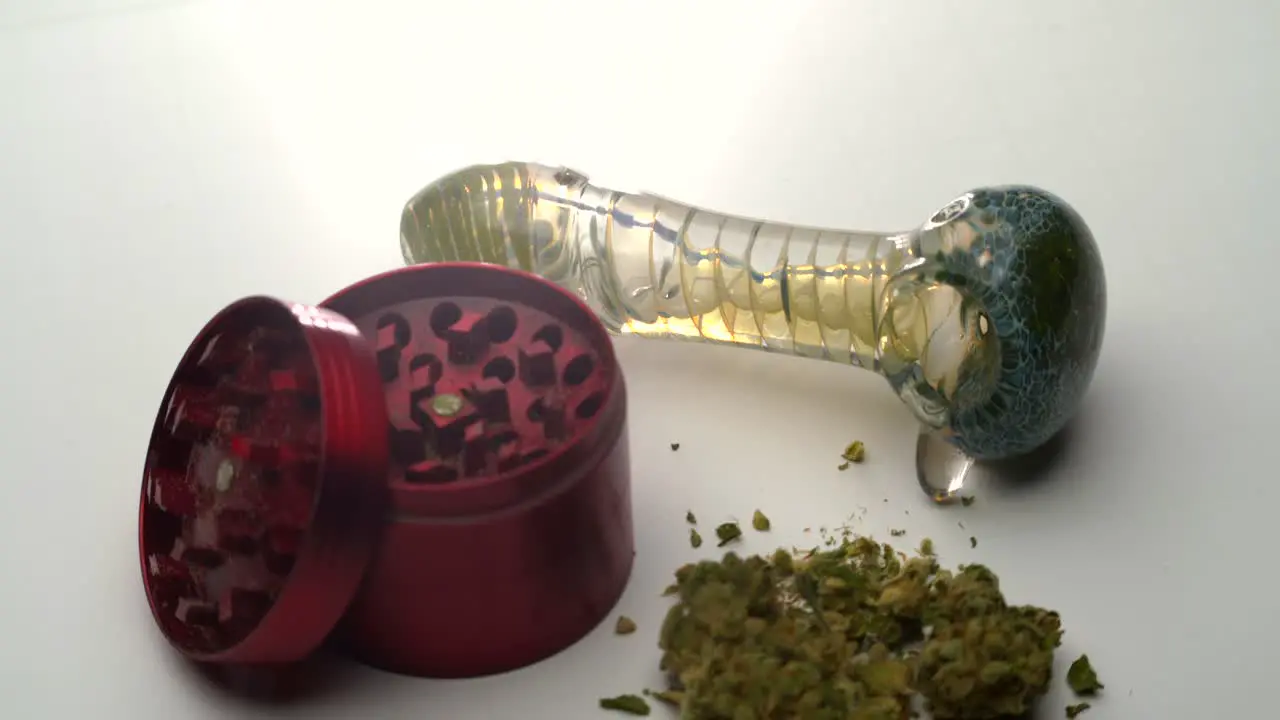 Closeup view of a glass pipe on a white table as the camera pulls back to reveal an open red grinder and marijuana buds