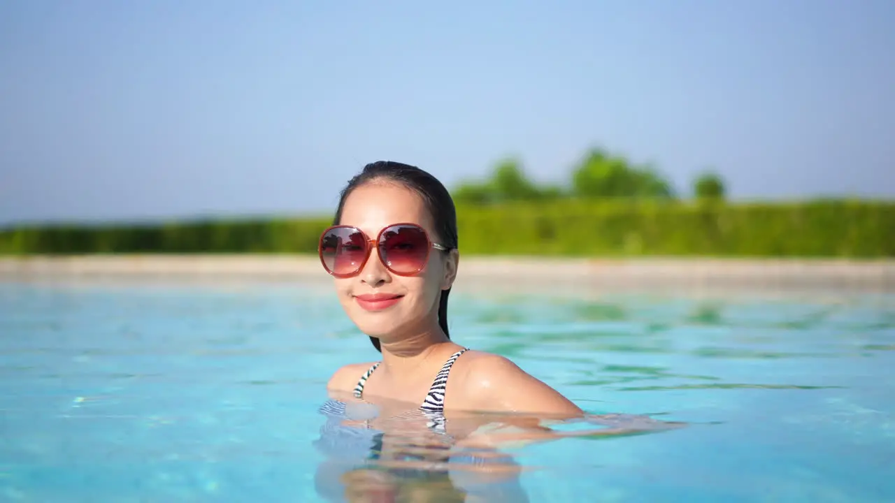 Sexy Asian woman with big sunglasses standing in swimming pool smiling to the camera