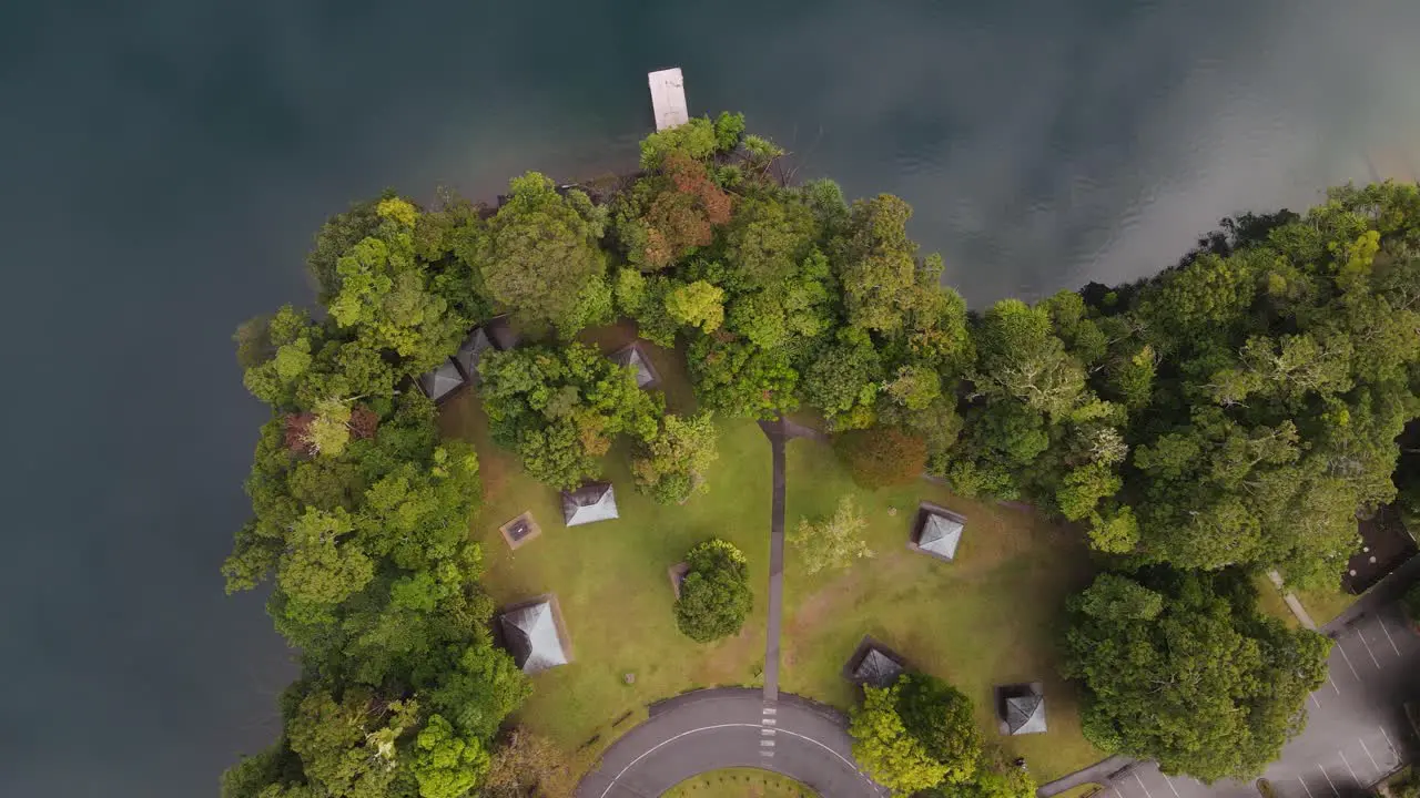 Moving drone video looking down on a lush park area next to a volcanic crater lake with clouds reflecting on the calm water