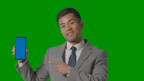 Studio Shot Of Smiling Businessman In Suit Holding Blue Screen Mobile Phone Against Green Screen 