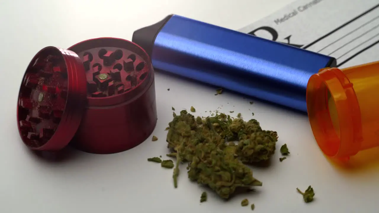 A small pile of ground marijuana lies on table surrounded by a grinder vaporizer open pill bottle and prescription note