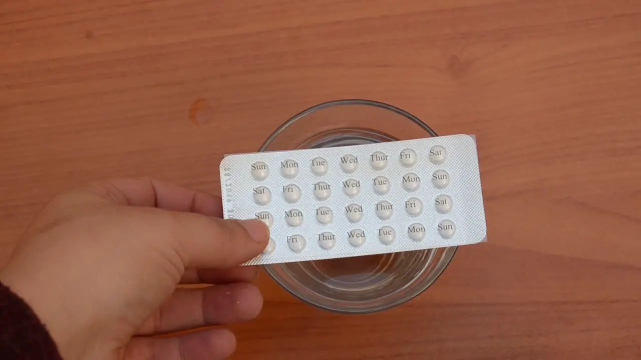 woman taking contraceptive pills from the top of the glass of water on the wooden table