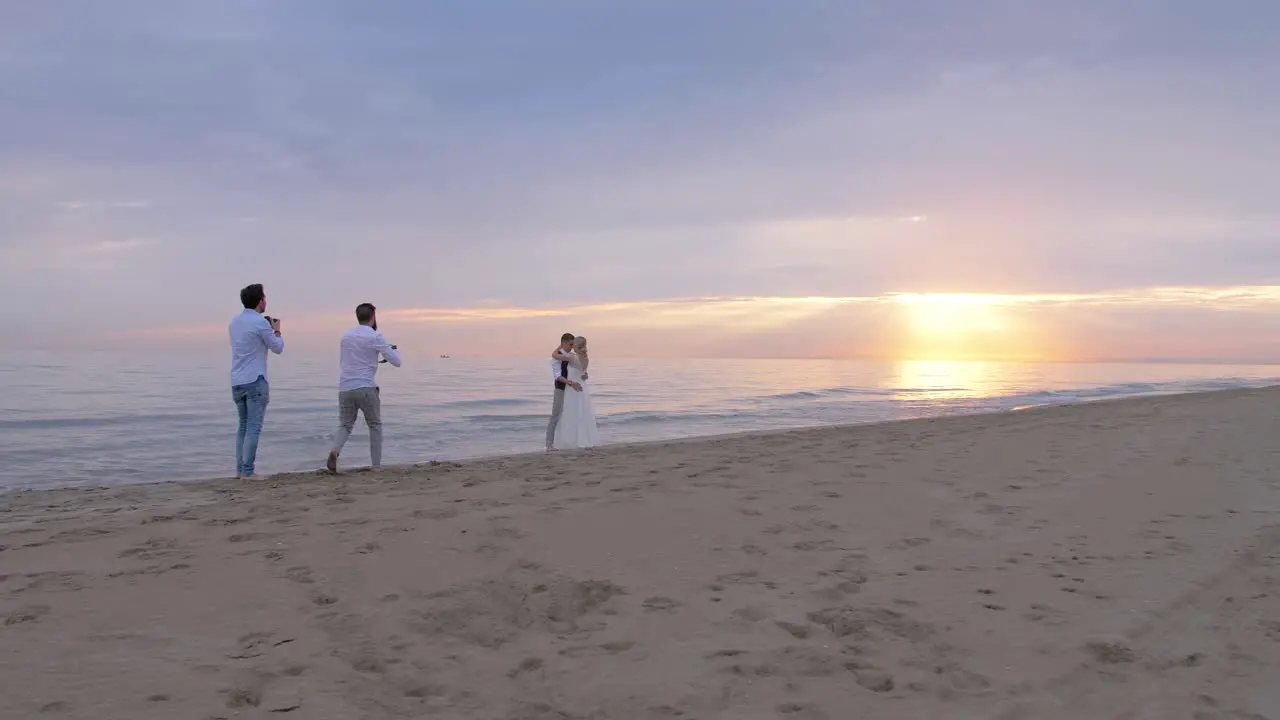 wide shot of the beach with the sun setting over the sea of two photographers shooting a newlywed couple during their wedding