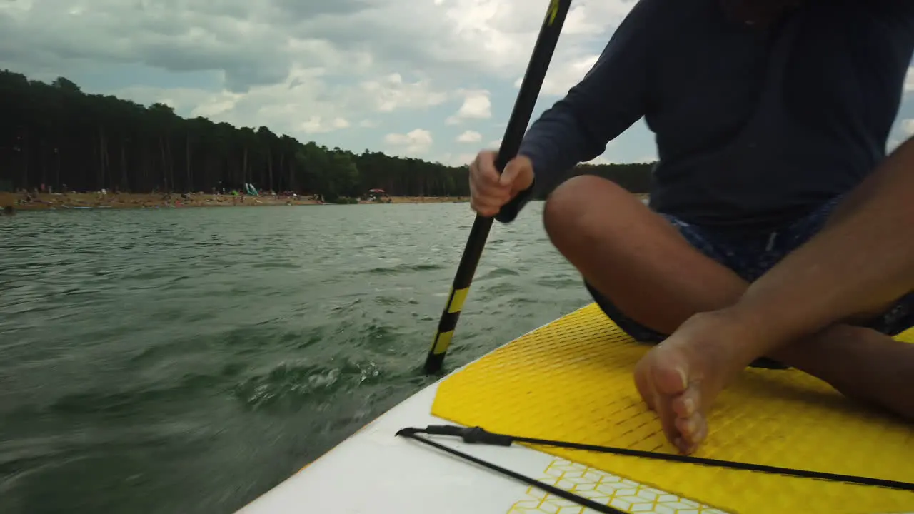 Man paddles fast while seated on paddleboard past lake beach low angle