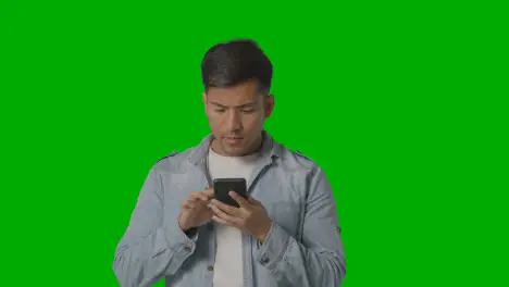 Studio Shot Of Casually Dressed Young Man Online Swiping On Mobile Phone Against Green Screen 