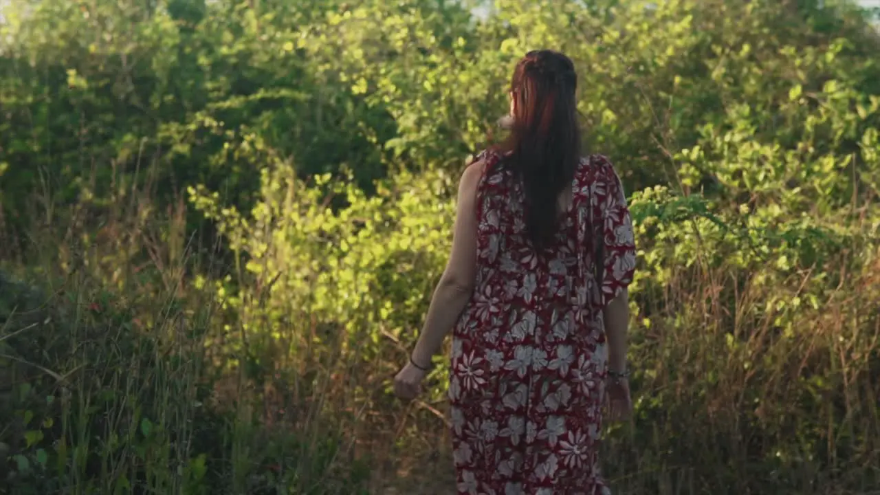 Out of focus brunette Indian woman walking away through high grass and trees wearing a red summer dress