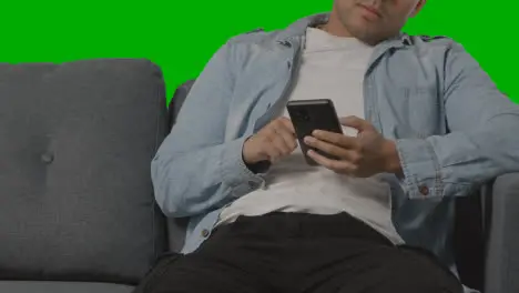 Close Up Of Bored Young Man Sitting On Sofa Looking At Mobile Phone Against Green Screen