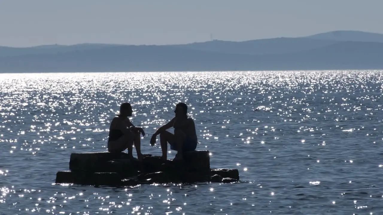 A couple sitting and chatting on a small rock formation in the middle of a shimmering Adriatic Sea