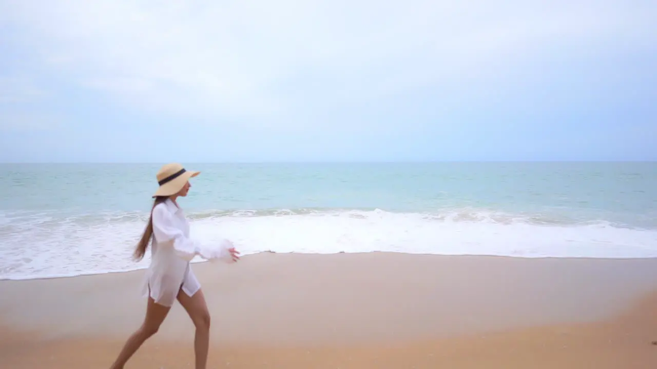 Slow-motion of a cute young woman in a white bathing suit walking across a sandy beach as the waves come in