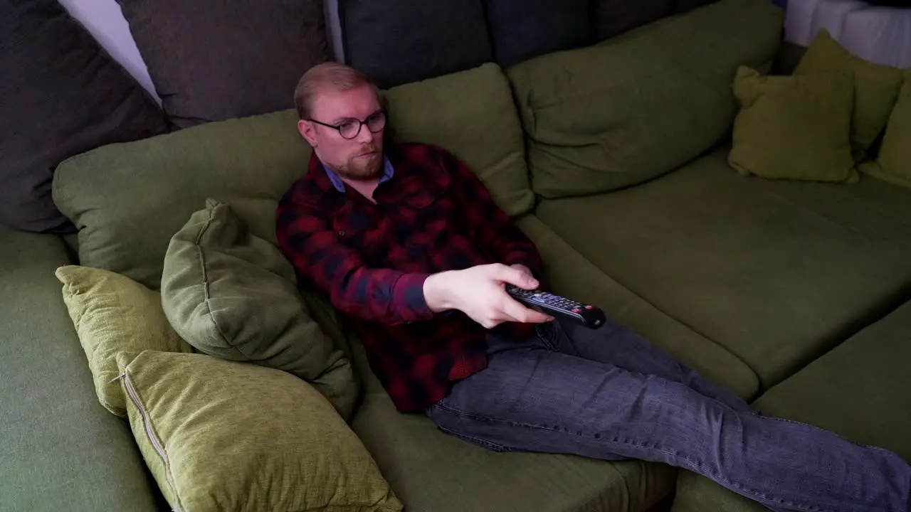 Young Man Lays on Sofa Uses TV Remote to Switch Channels Medium Shot