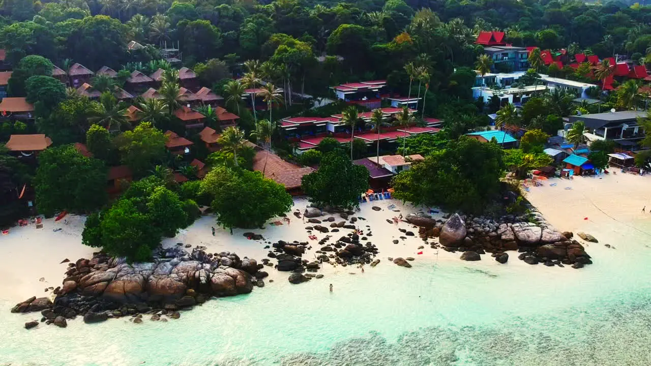 An aerial shot of a resorts by the beach with few unrecognisable people walking along the shoreline and trees in the background