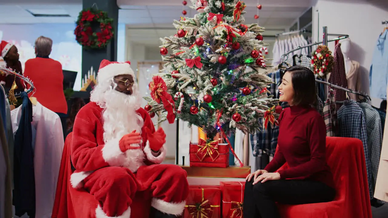 Dolly out shot of worker acting as Santa Claus sitting down next to Christmas tree with asian woman in xmas decorated clothing store Fashion shop retail assistant offering present to customer