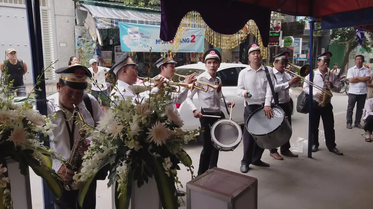 A brass band dressed in French Colonial uniform play loud music an important ritual at a funeral in a narrow urban street in Vietnam