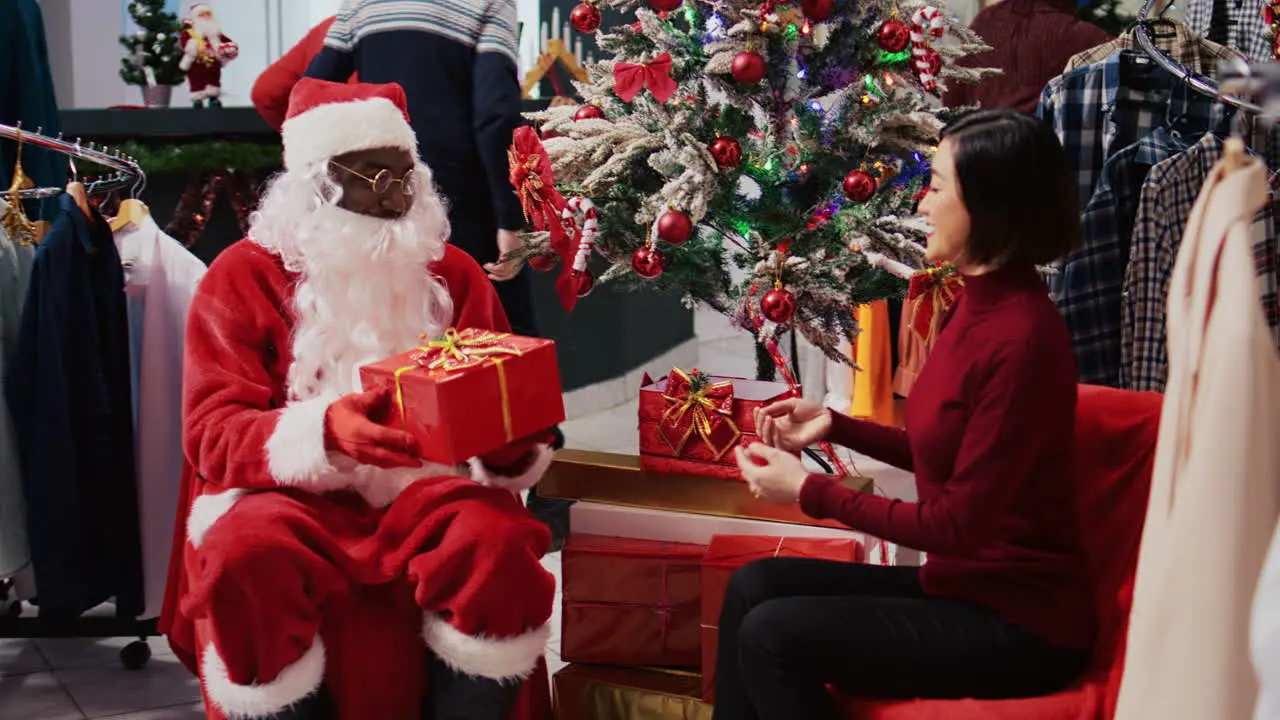 African american manager acting as Santa Claus sitting down next to Xmas tree with woman in festive decorated clothing store Fashion boutique supervisor offering present to grateful client