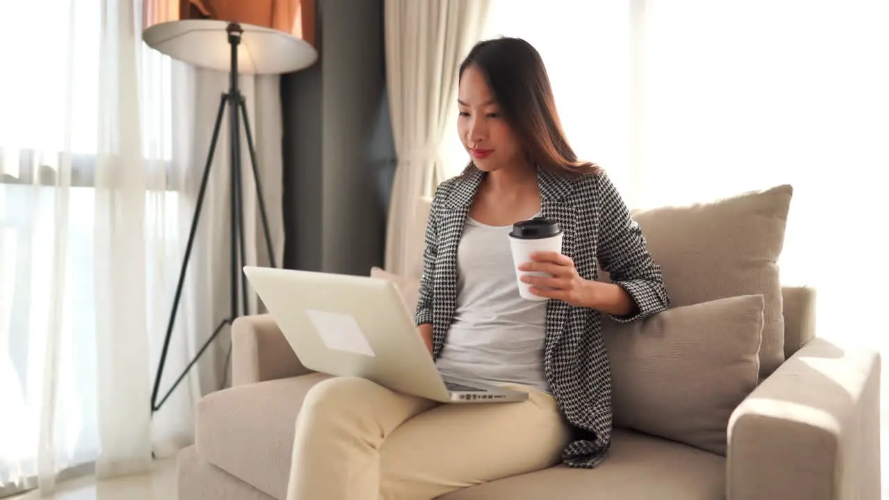 A pretty Asian woman sitting on an overstuffed chair holding a coffee cup while typing on her laptop