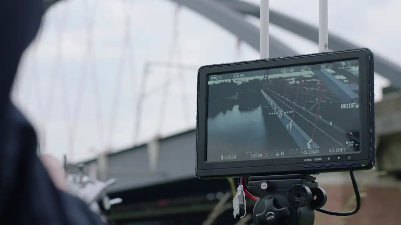 View from handheld camera of operator focusing tripod-mounted display on blue-arch bridge with red cables with settings visible on connected monitor