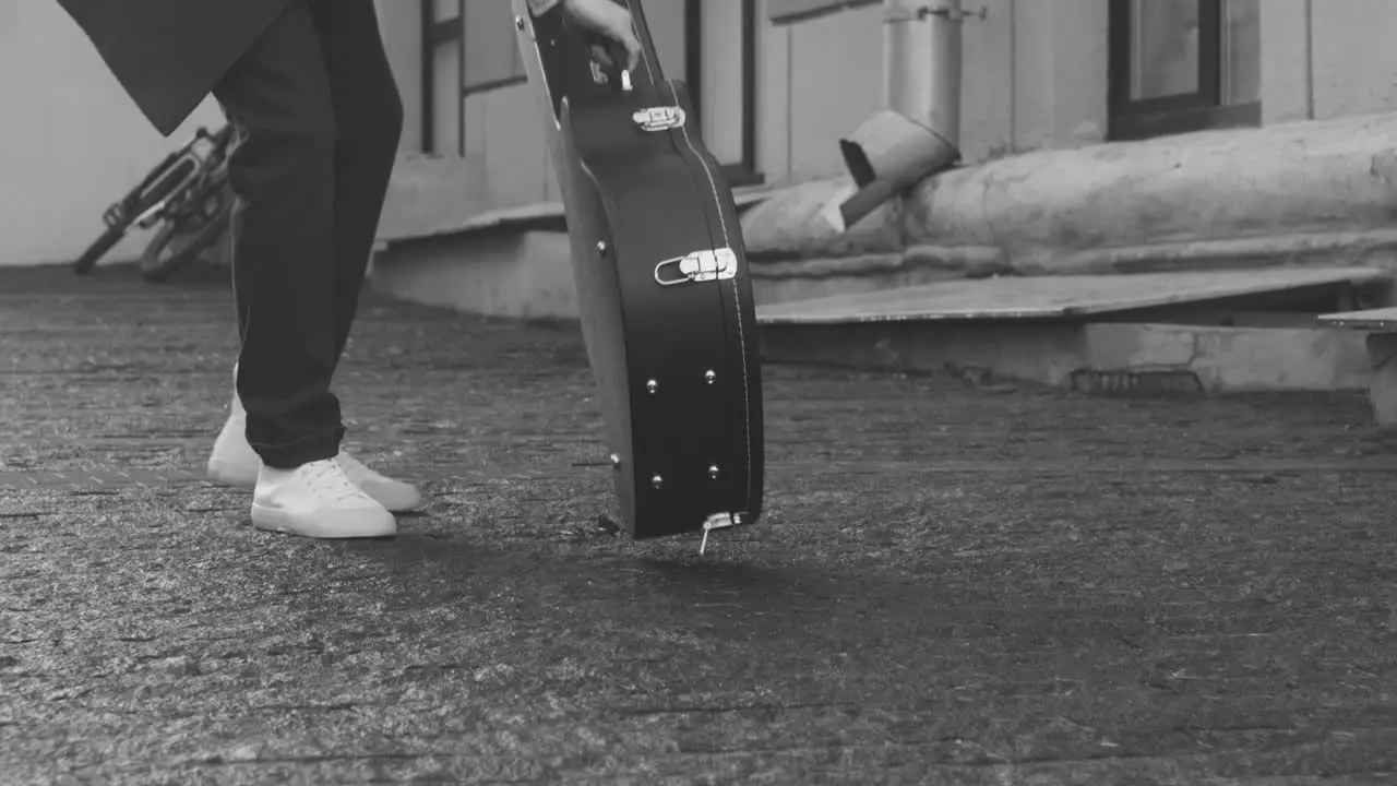 Black And White View Of A Man In Coat Picking Guitar From A Case On The Floor Outdoors