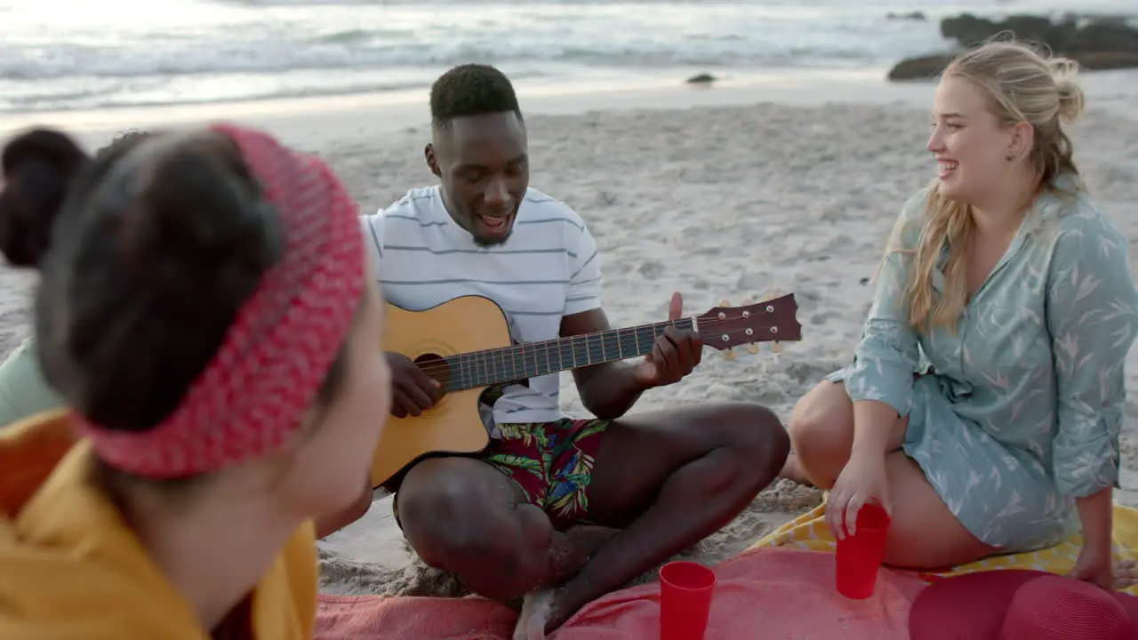 Young African American man plays guitar on the beach at a party surrounded by friends