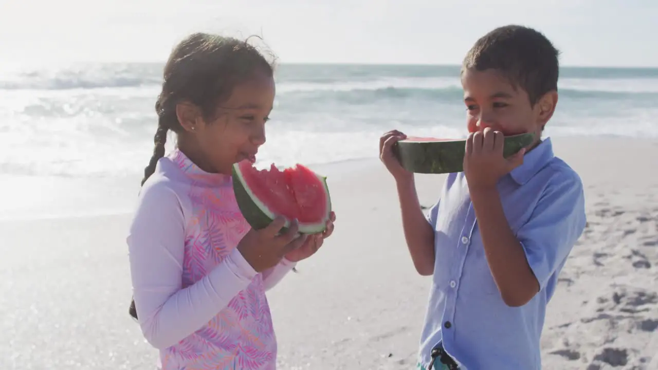 Happy hispanic brother and sister eating watermelon on beach