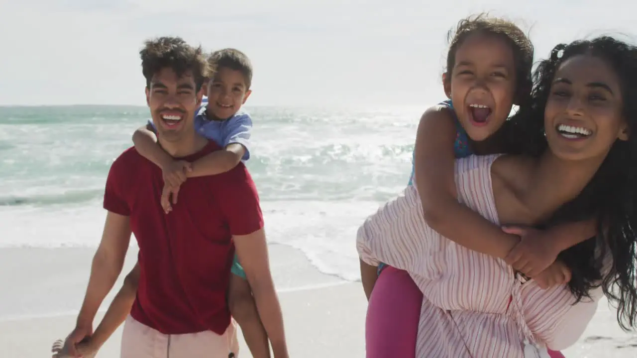 Happy hispanic parents carrying children on piggyback on beach