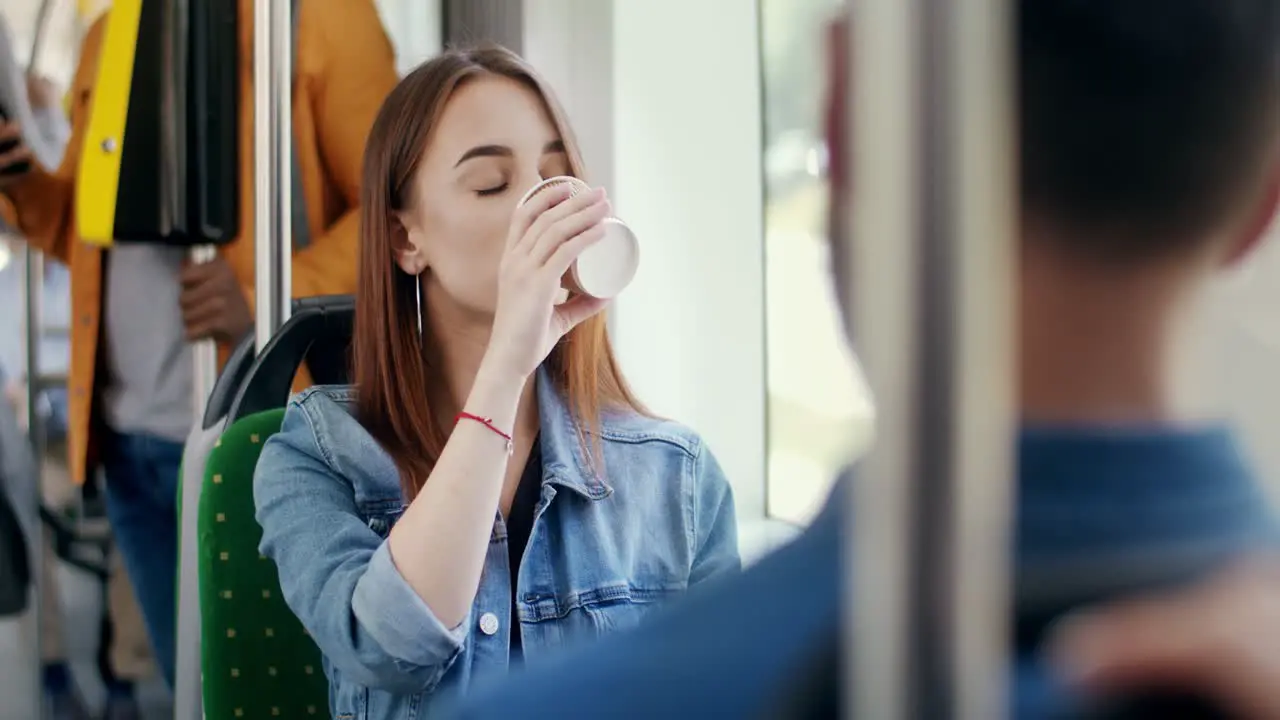 Young Beautiful Girl Sitting In The Tram While Going Somewhere Holding A Cup To Go And Sipping Hot Coffee While Smiling And Flirting To The Guy In Front Of Her