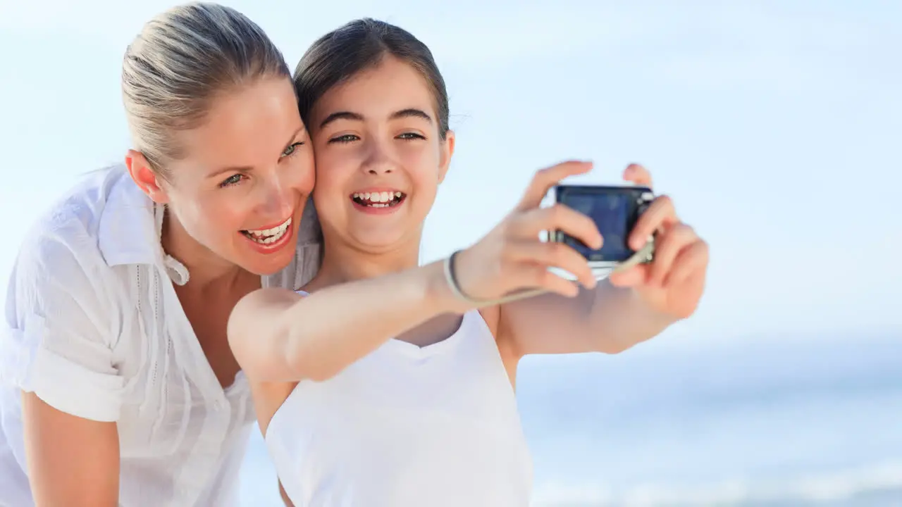 Portrait of smiling caucasian mother and daughter on holiday taking photos by sea