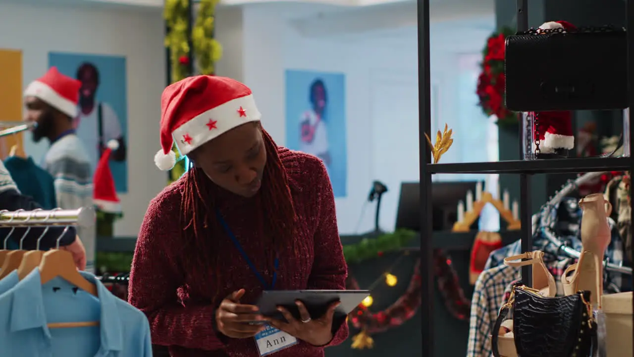 Employee wearing festive Santa hat crosschecking online shop clothing prices with physical store ones looking to correct mismatches Worker in Christmas decorated fashion boutique