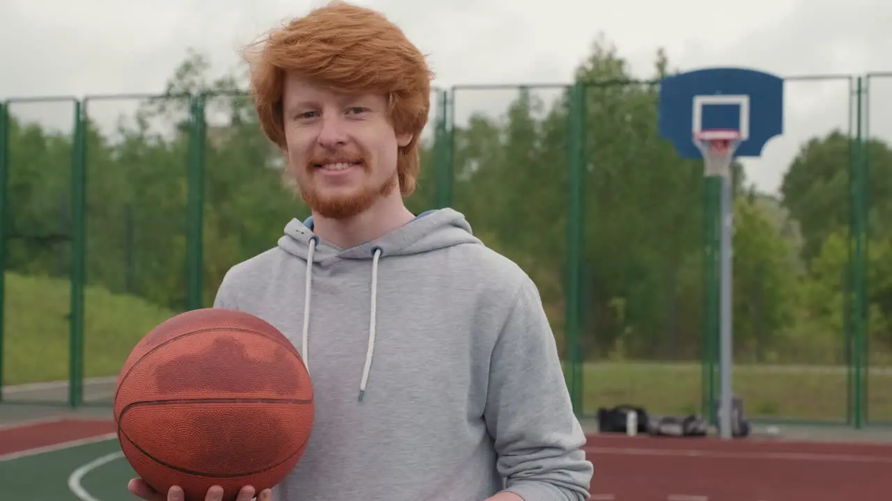 Young Smiling Redhead Athlete With Sweatshirt Holding Ball For Playing Basketball In Basketball Court