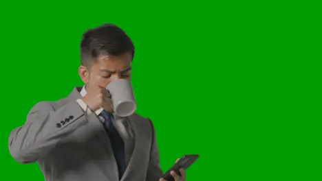 Studio Shot Of Businessman In Suit Holding Hot Drink Looking At Mobile Phone Against Green Screen 