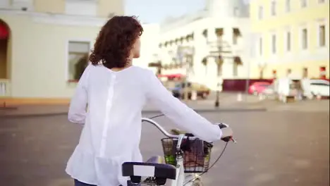 Back view of a woman in a white shirt and blue jeans walking in the city street holding her bicycle handlebar with a basket