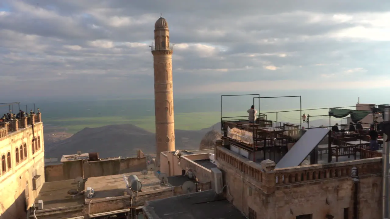 As the camera pans to the right we see the roof cafes of Mardin and the minaret of the Great Mosque behind them and the magnificent view of Mesopotamia at the back