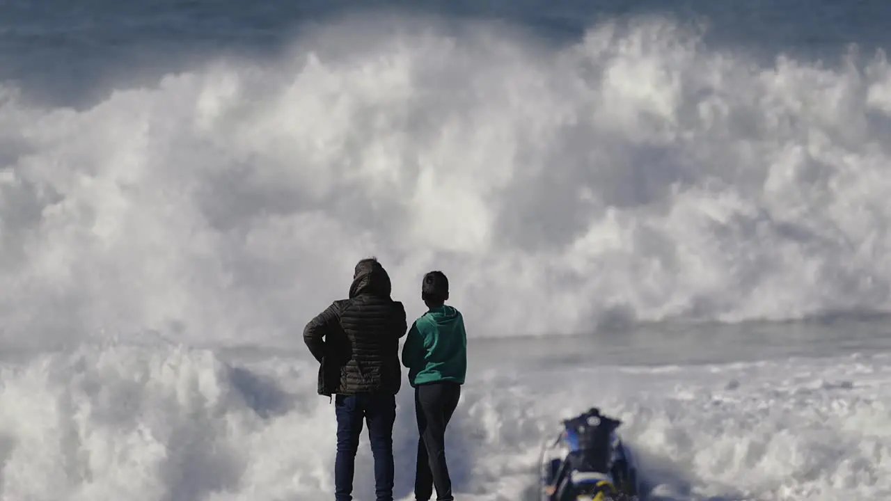 Two people watch a jet ski driver from the beach as he rides a foamy wave that throws him high