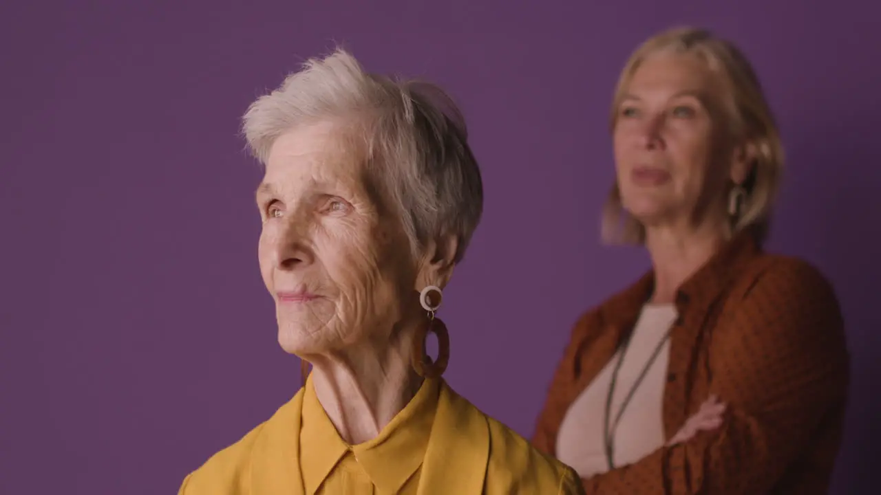 Blonde Senior Woman With Short Hair Wearing Mustard Colored Shirt And Jacket And Earrings Posing With Blurred Mature Woman On Purple Background 1