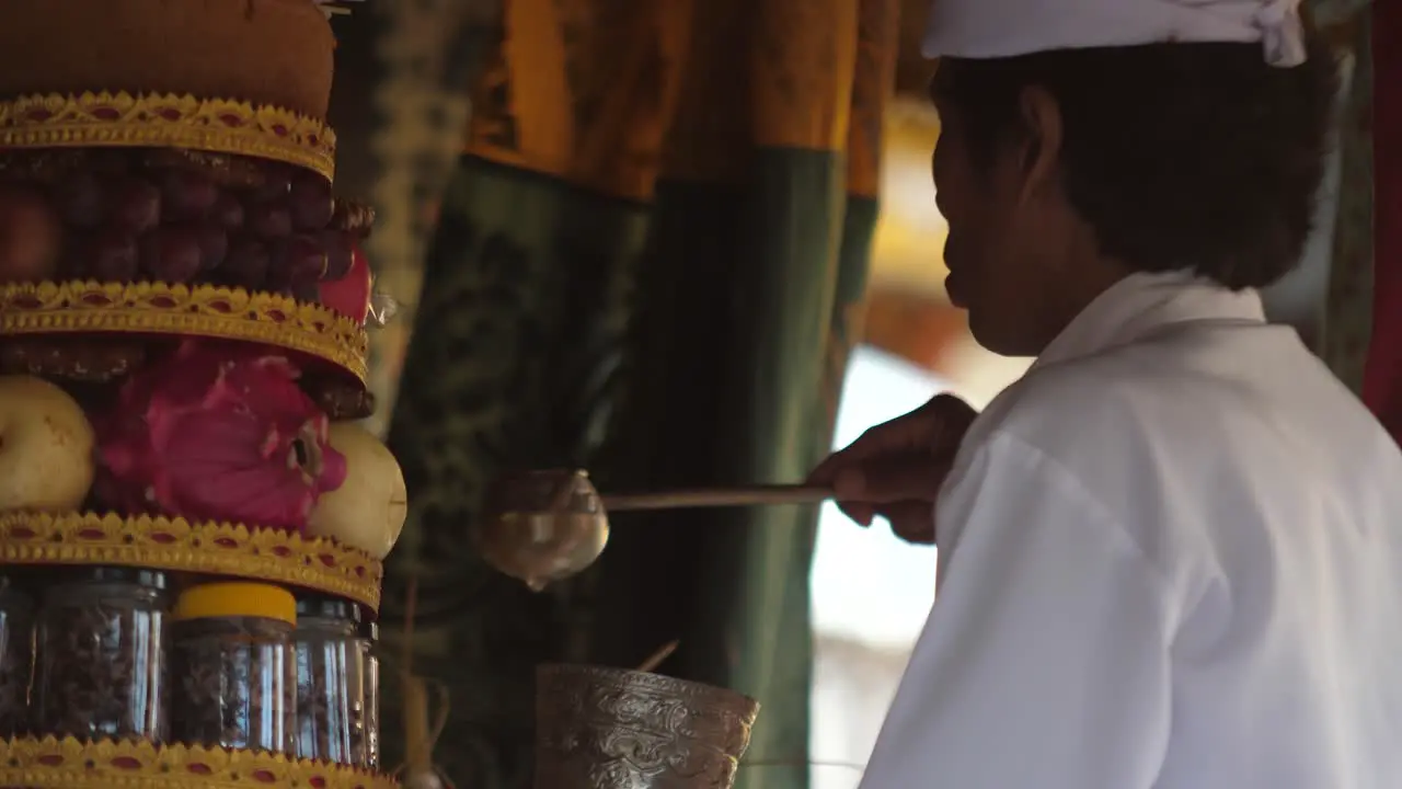 Food offerings on altar at Hindu temple in Bali Indonesia as priest in white clothing scoops water into silver cup for blessing and ceremony