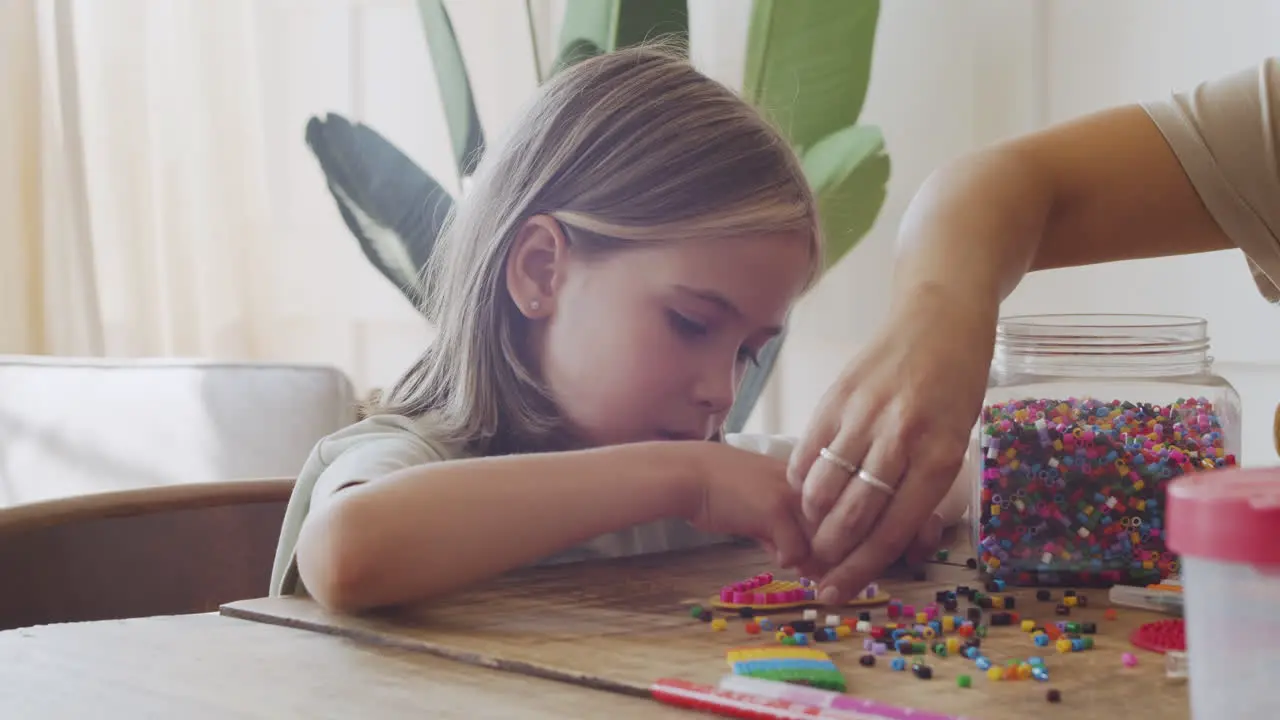 Close Up Of A Cute Little Blonde Girl Having Fun Playing With Colored Beads Helped By Her Mother