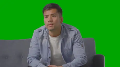 Studio Shot Of Tense Young Man Sitting On Sofa Watching Sport On TV Against Green Screen 