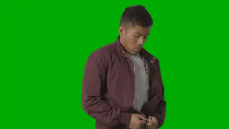 Portrait Of Cold Shivering Casually Dressed Young Man Doing Up Coat Against Green Screen 