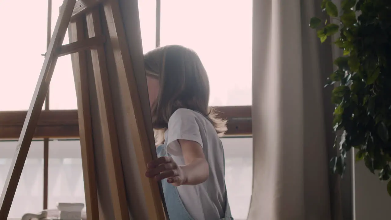 Little Blonde Girl Painting On The Lectern Next To The Window At Home