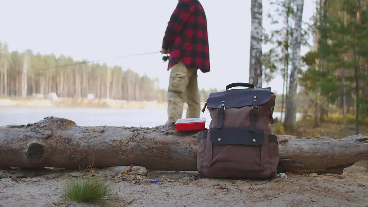 man is fishing in lake standing on coast in forest backpack with equipment and baits on groundrelax hobby