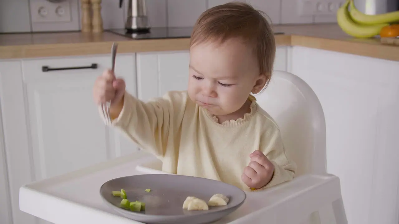 Cute Baby Girl Sitting In High Chair In The Kitchen And Holding Fork And Eating Fruit Slices