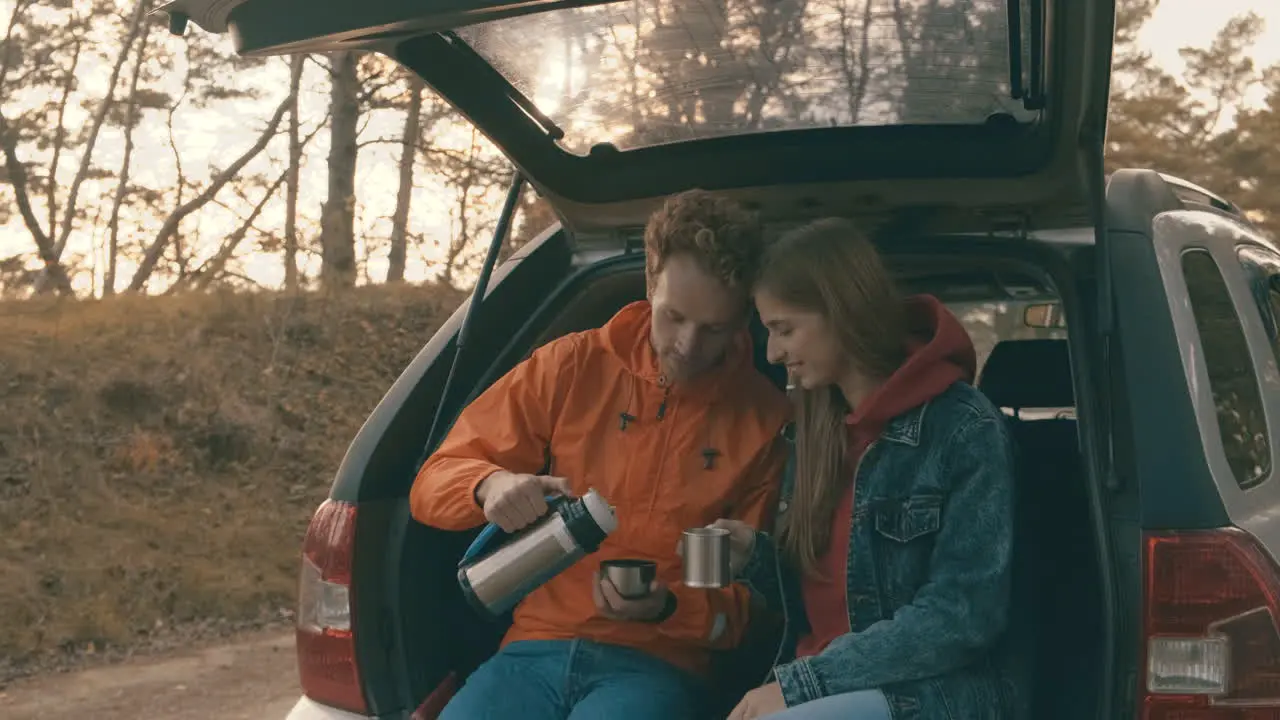  Young woman and man sitting in the open trunk of the car having a hot drink with a thermos