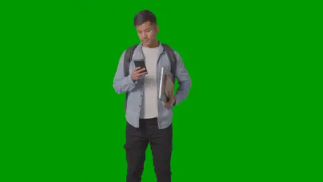 Three Quarter Length Shot Of Male University Or College Student Looking At Mobile Phone Against Green Screen 