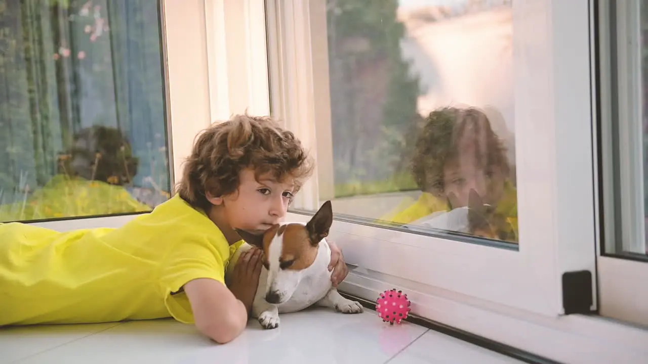 Blonder Junge Mit Lockigen Haaren Der Mit Seinem Hund Auf Dem Boden Liegt Ihn Streichelt Und Aus Dem Fenster Schaut 1