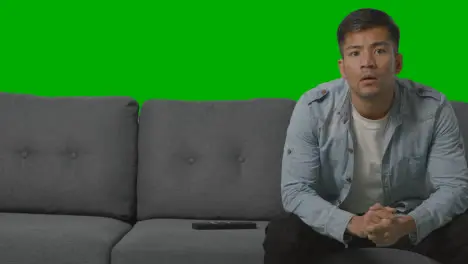 Studio Shot Of Disappointed Young Man Sitting On Sofa Watching Sport On TV Against Green Screen 