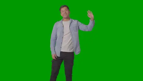 Portrait Of Casually Dressed Smiling Young Man Waving In Welcome Against Green Screen 