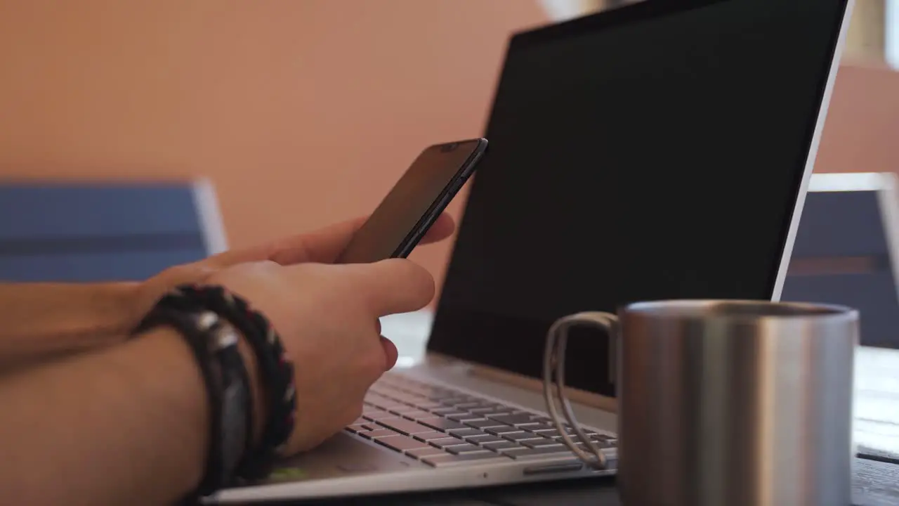 Closeup of hands typing on the mobile phone with a steel mug and laptop in the background