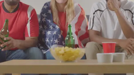 Multi-Cultural Friends Supporting USA Team Watching Sports Game On TV Sitting On Sofa At Home With Flag