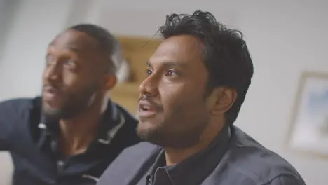 Two Male Friends Celebrating Watching Sports Game On TV Sitting On Sofa At Home Drinking Beer