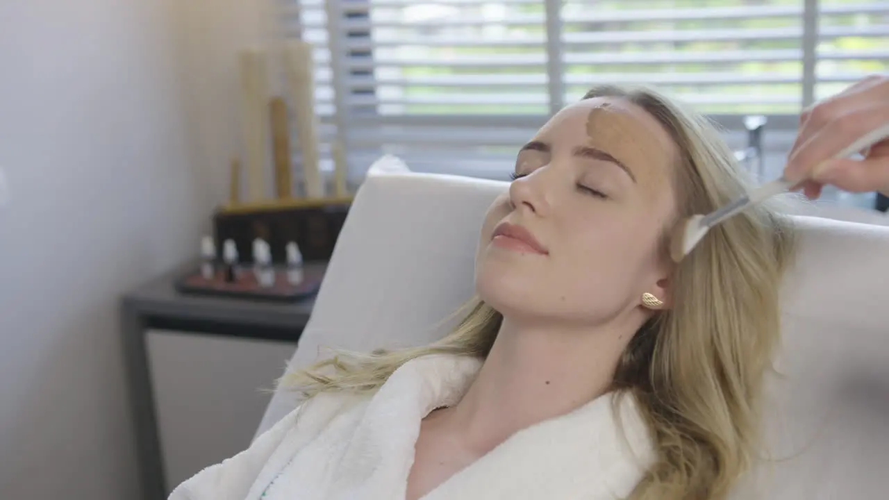 Young blonde caucasian woman receive a health skin care facial treatment while lying on a massage bed in luxury resort spa saloon