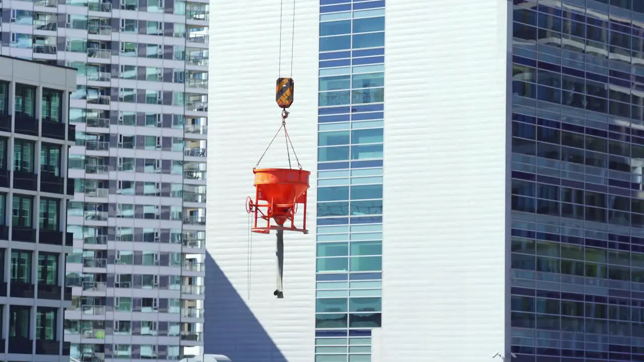 Orange bucket with concrete hung from crane in front of new buildings with windows